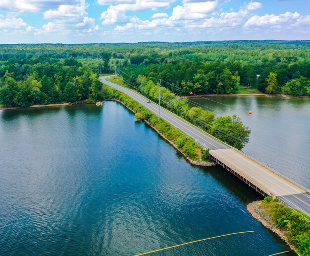 Aerial shot of a bridge, road, trees near the lake with a cloudy blue sky