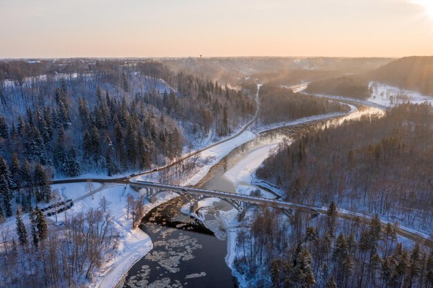 Aerial shot of a curvy road over a flowing river through the snow-covered forest