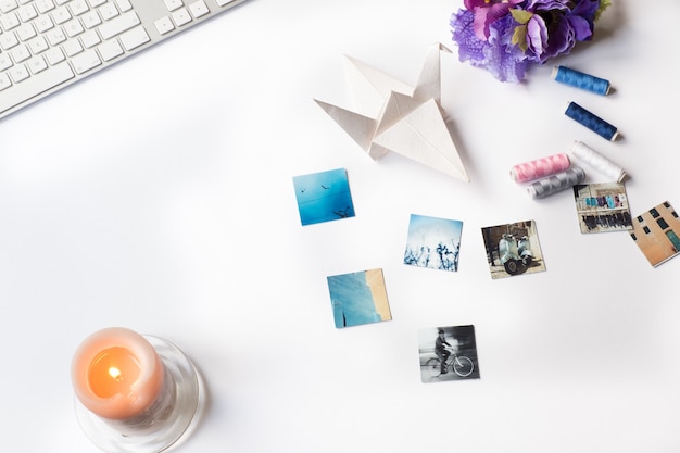 Aerial shot of photos, a slim keyboard, an orange candle, paper origami and thread on a white desk