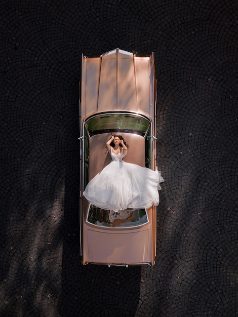 Aerial view of elegant bride lying on the roof of a pink retro car