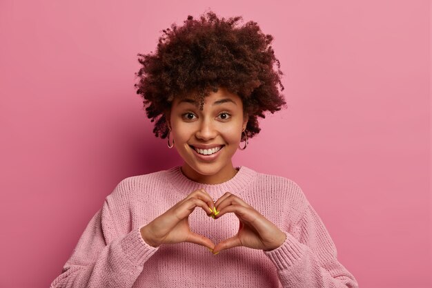 Affection and relationship concept. Glad ethnic delighted woman shapes heart with hands, smiles positively, demonstrates love symbol, wears pink pastel sweater, gestures indoor. Monochrome shot