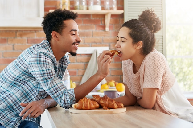 Affectionate bearded mixed race man feeds his girlfriend with tasty croissant which he baked by himself.
