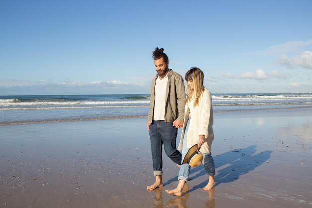 Ласковая пара гуляет у воды в летний день. Бородатый мужчина и женщина в повседневной одежде держатся за руки, прогуливаясь по берегу моря. Концепция любви, путешествий, свиданий