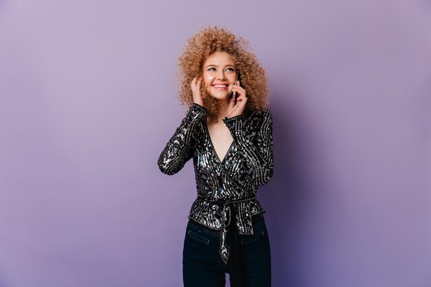 Affectionate curly girl dressed in shiny bright top and dark denim pants with smile talking on phone on isolated background