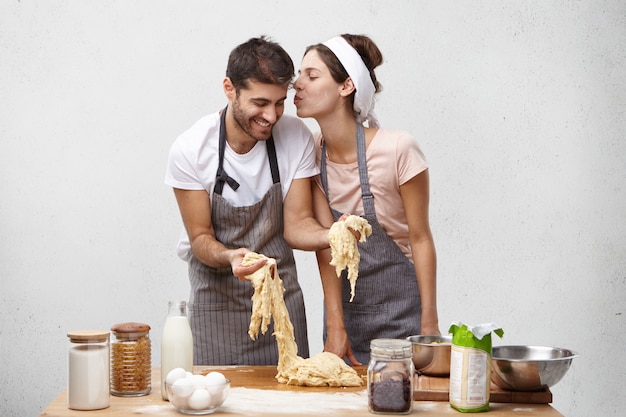 Affectionate female going to kiss hard working husband who makes dough and helps her at kitchen