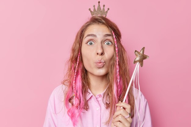 Affectionate tender woman with long hair keeps lips rounded expresses love to someone poses with magical wand makes wish wears little crown isolated over pink background Lovely queen indoor