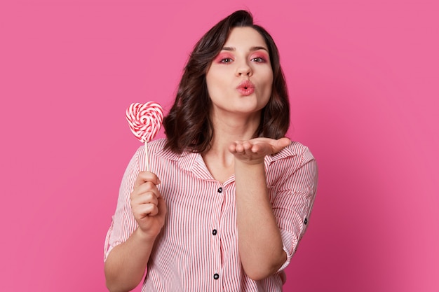 affectionate woman blows air kiss, dressed in fashionable clothes, holds candy, has bright makeup, poses on rosy. Its love is for you. Lovely pinup girl has fun indoor.