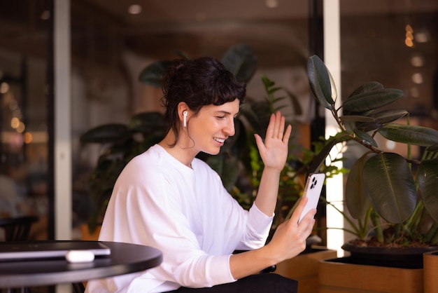 Affectionate young caucasian brunette woman in casual clothes waving at smartphone screen sitting indoors