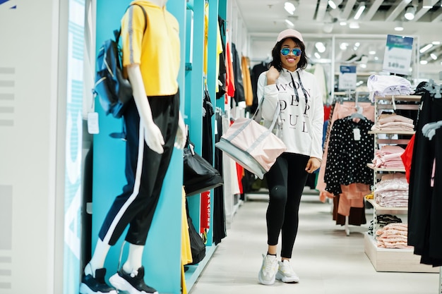 운동복과 선글라스를 착용한 아프리카계 미국인 여성들은 선반에 스포츠 가방이 달린 스포츠웨어 쇼핑몰에서 쇼핑합니다. 스포츠 스토어 테마