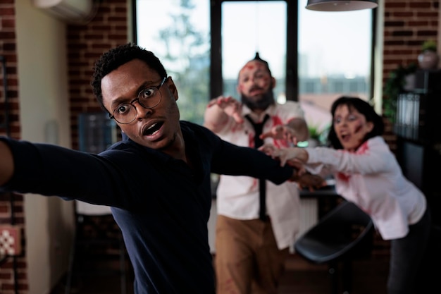 Afraid man running from scary zombies, trying to eat human brain in business startup office. Bloodthirsty eerie monsters with wounds chasing after frightened scared person, angry corpses.