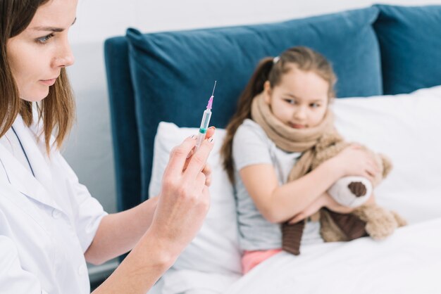 Afraid sick girl looking at female doctor hands filling the syringe with medicine