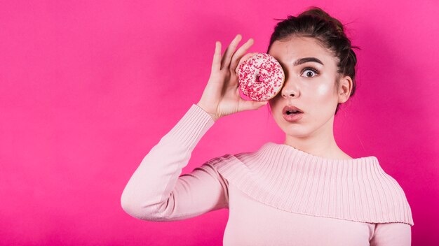 Испуганная молодая женщина держит пончик на глазах на розовом фоне