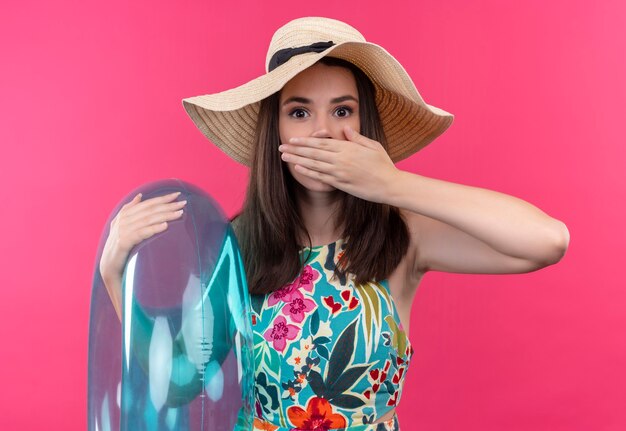 Afraid young woman wearing hat holding swim ring and holding hand on her mouth on isolated pink wall