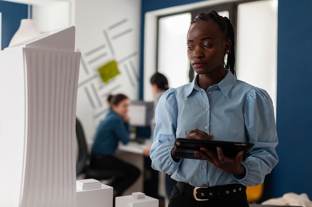 African america architect looking at scale foam model of modern building while holding tablet with blueprints. Architectural engineer focused on the design of 3d skyscraper maquette.