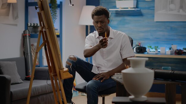 African american artist looking at vase design for inspiration to draw on canvas in creativity studio room at home. Black adult man using pencil for masterpiece drawing, creating fine art