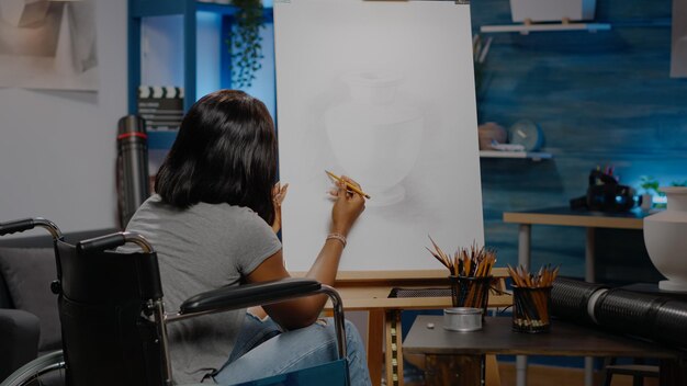 アートワーク空間での専門的な職業のための花瓶のデザインを描く障害を持つアフリカ系アメリカ人の芸術家。革新的な傑作に取り組んでいる間車椅子に座っている黒人の若い女性