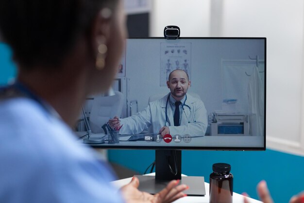 病院のオフィスで働くオンラインビデオ通話会議中に、遠隔地の医師とインフルエンザの症状について話し合うアフリカ系アメリカ人のアシスタント。コンピュータ画面上の遠隔医療呼び出し
