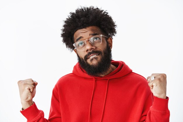 african american bearded guy in glasses biting lower lip in desire and faith clenching fists hoping hear positive news daydreaming about victory on gray wall.