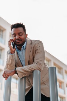 Афро-американский мальчик в костюме разговаривает по смартфону с клиентом своей компании в месте, где видны здания компании. копировать пространство.
