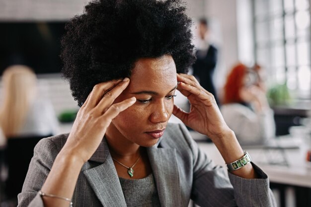 Афроамериканская деловая женщина, у которой болит голова и она чувствует себя недовольной из-за проблем на работе. На заднем плане люди.