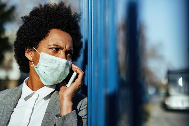 African American businesswoman with face mask talking on cell phone while being outdoors at bus station