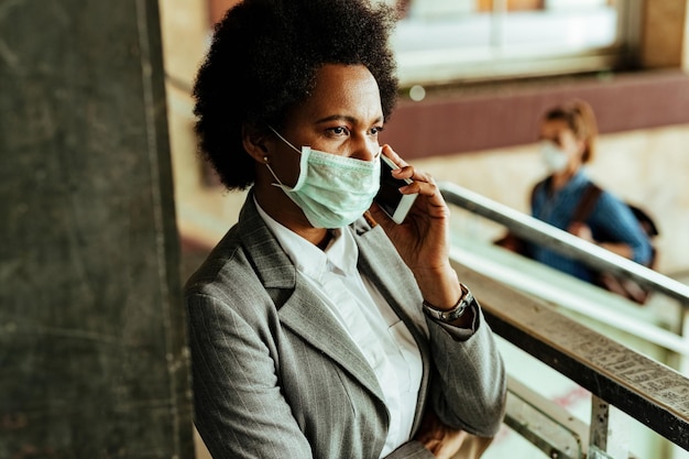 African American businesswoman with protective face mask communicating over smart phone while being at public hallway at railroad station