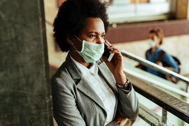 Афроамериканская деловая женщина в защитной маске общается по смартфону, находясь в общественном коридоре на вокзале