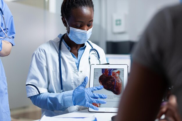 African american cardiologist doctor showing heart radiohraphy to sick patient using tablet explaining medication treatment during clinical appointment in hospital office. People with medical face mas