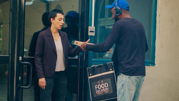 アフリカ系アメリカ人の運送業者がドアベルを鳴らしてファーストフードの食事の注文を届け、持ち帰り用のバックパックを運ぶ.オフィス ビルの顧客にランチ食品を提供するレストランの宅配便。手持ち撮影。