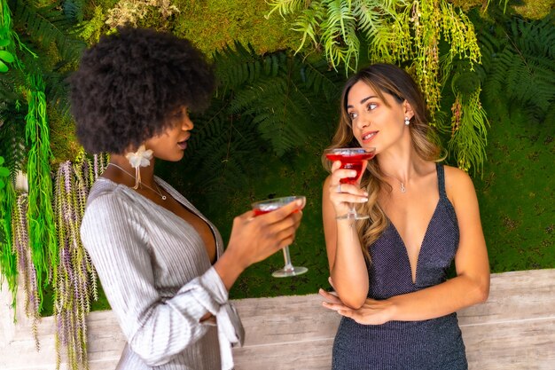 African-American and Caucasian female friends wearing fancy dresses drinking wine and talking