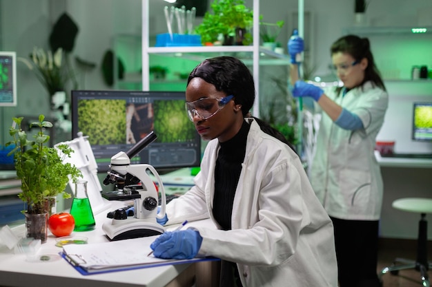 African american chemist woman writing medicine experiment results on clipboard after analyzing genetic mutation on plant sample under microscope. Scientist working in biochemistry laboratory