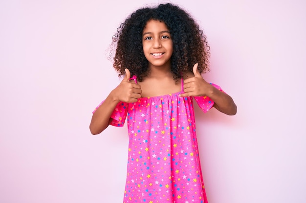 Free photo african american child with curly hair wearing casual dress success sign doing positive gesture with hand, thumbs up smiling and happy. cheerful expression and winner gesture.