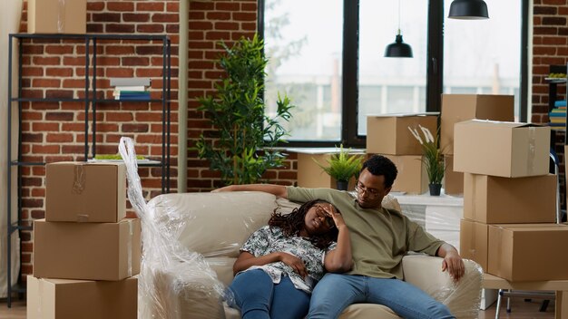 African american couple unpacking couch and furniture to decorate new rented apartment flat. Taking break from moving boxes and belongings, buying first household and celebrating life event.