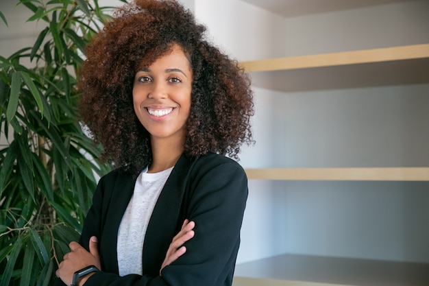 African American curly businesswoman standing with folded hands. Portrait of successful confident young pretty female office employer in suit posing at work. Business, company and management concept