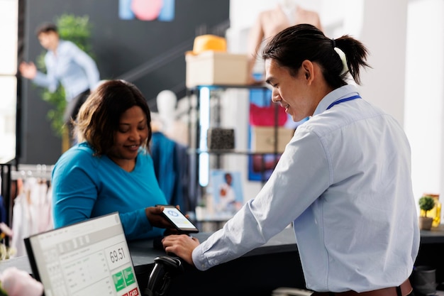 African american customer putting phone on pos terminal paying for stylish clothes in boutique after discussing items fabric with employee. Woman standing at retail store counter buying casual wear