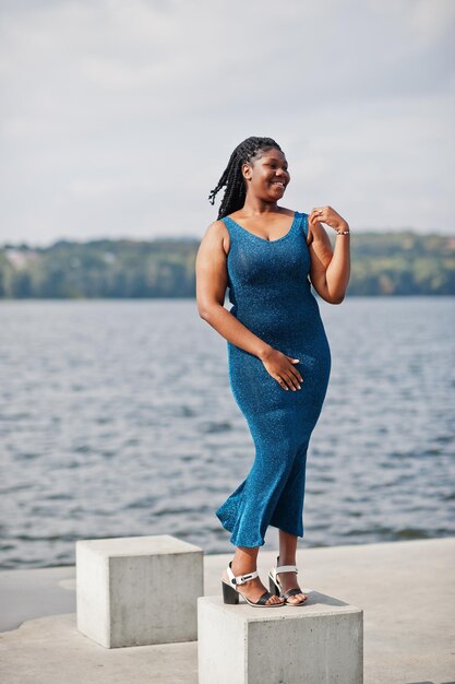 아프리카계 미국인 검은 피부 플러스 사이즈 모델은 바다 쪽을 배경으로 파란색 반짝이는 드레스를 입고 포즈를 취했습니다.