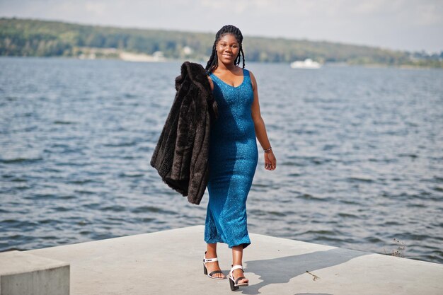 African american dark skinned plus size model posed in a blue shiny dress and black fur coat against sea side