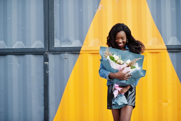 African american dark skinned slim model posed in a black leather shorts and jeans jacket She hold bouquet of flowers against yellow triangle steel wall