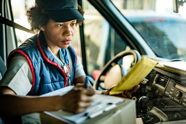 Foto gratuita donna di consegna afroamericana che controlla l'indirizzo su un pacco e prende appunti mentre è seduto in un furgone