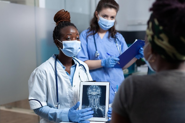 African american doctor explaining medical radiography to sick patient discussing healthcare treatment with nurse during clinical examination in hospital office. people with face mask against covid19