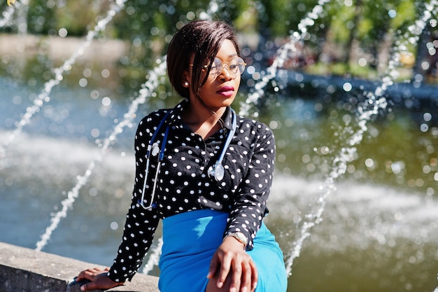 聴診器を持つアフリカ系アメリカ人の医師の女性は、屋外の背景の噴水を提起しました