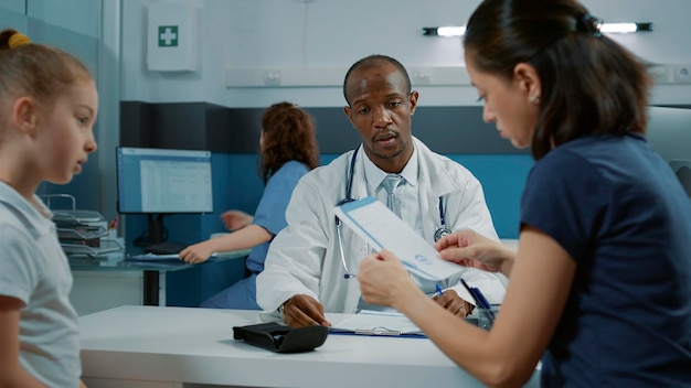 African american doctor giving prescription medicine paper to parent in cabinet at checkup visit. Mother of little girl receiving medical treatment and cure from general practitioner at clinic.