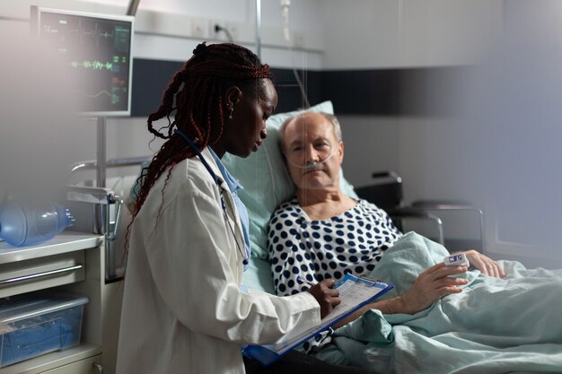 African american doctor in hospital room discussing diagnosis and treatment with sick senior man lay...