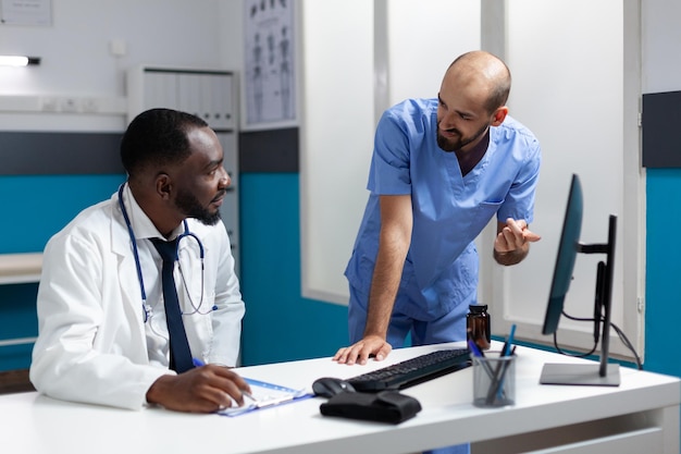 African american doctor monitoring sickness expertise on computer discussing healthcare treatment with man nurse. Medical team of physician working at medicine prescription in hospital office