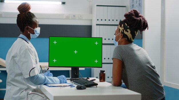 African american doctor and patient looking at green screen on computer in cabinet. Black medic and woman watching monitor with mockup template and isolated background for chroma key.