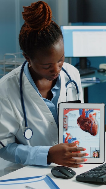 Афро-американский врач показывает пациенту сердечно-сосудистое изображение на планшете для диагностики кардиологии. Черный медик объясняет молодому человеку состояние сердца и кровоток с помощью гаджета