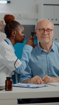 耳鏡​を​使用している​アフリカ系​アメリカ人​の​医師​は​、​医療​キャビネット​で​病気​の​老人​に​相談します​。​机​の​上​の​年長​の​患者​に​プロ​の​ツール​で​耳​の​検査​を​している​黒人​の​耳科医