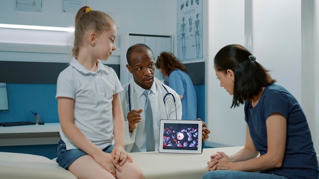 African american doctor using tablet to show virus illustration. Specialist explaining virus animation on display to mother and child at medical consultation in cabinet. Health care visit