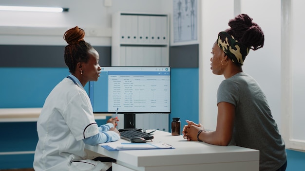 予定と医療制度に関する情報のためにコンピューター画面を見ているアフリカ系アメリカ人の医師と女性。毎年の健康診断のために薬を持って机に座っている患者。