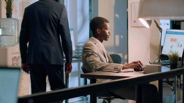 Free photo african american employee crosschecking financial figures between clipboard charts and diagrams on computer screen during nightshift efficient worker comparing company numbers in office